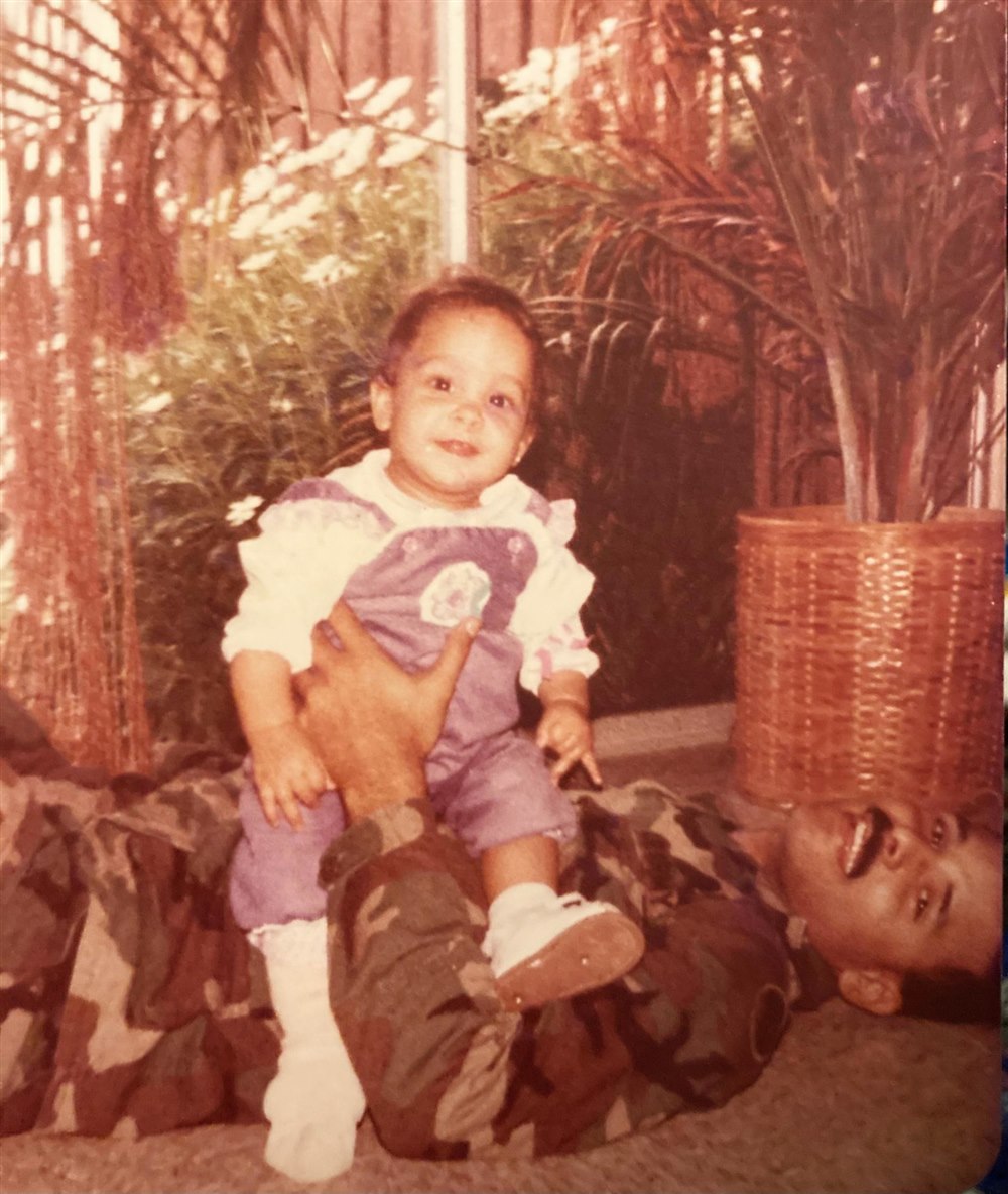 Baby Alani sitting on her father, who is lying down in his military camouflage uniform