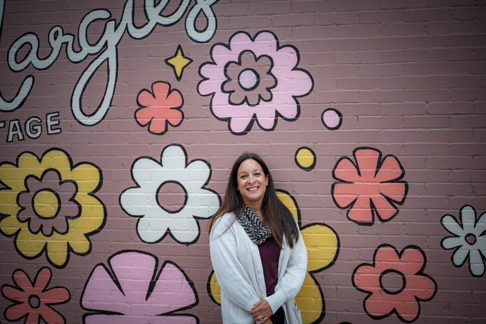 Alani Bankhead standing in front of a pink brick wall painted with large flowers, wide frame.