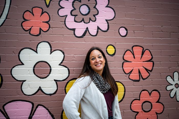 Alani Bankhead smiling in front of a pink brick wall painted with large flowers.